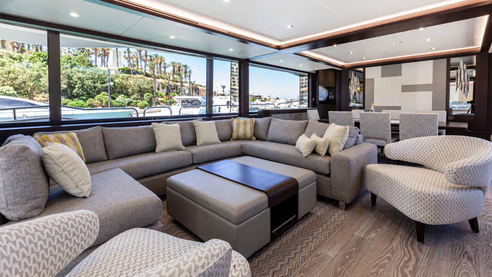 Daylit yacht lounge with grey sofas, large windows and palm trees outside
